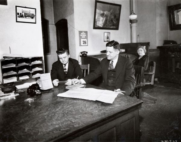 W.R. Boquet and A.W. Boquet sit together at a desk in the On Time Transfer Company office while Mrs. Erma Boquet looks on from the background.