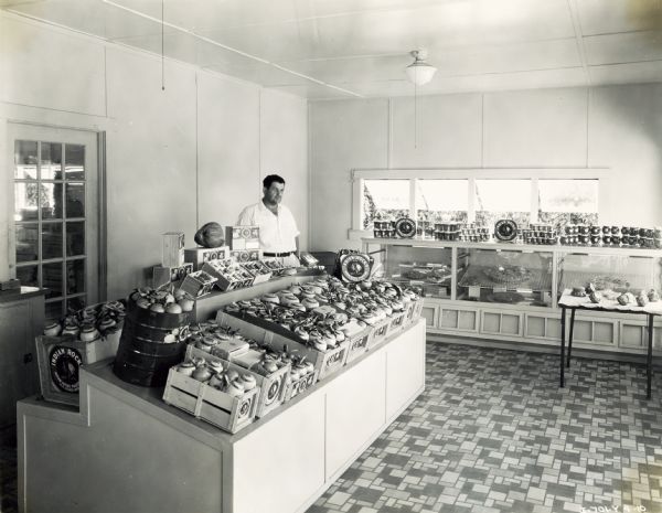 H.D. Ulmer stands behind the counter of his Indian Rocks brand fruit basket gift shop. The original caption reads: "H.D. Ulmer, owner of 75-acre farm near Largo,deals exclusively in gift boxes of selected fruit (Indian Rocks Brand). Mr. Ulmer owns PD-40 and P-30 power units operated in sawmills; also owns F-20 tractor used for snaking logs to sawmill. Operates an O-12 with a 7-foot 10-A disk harrow in his groves."