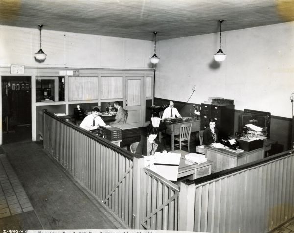 Men and women sit behind desks in the offices of Quinn R. Barton, Inc., an International Harvester dealership. Pictured in the photograph are: A.R. Zigler, secretary and treasurer; Troy Haigler, collector; Carolyn Marable, bookkeeper and stenographer; D.H. Sauls, bookkeeper; and Edith Berk, stenographer.