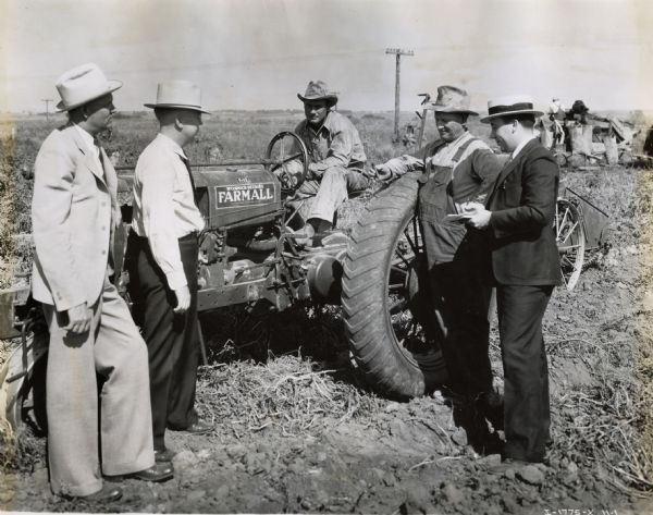 L.R. Love sits behind the wheel of a Farmall F-12 tractor pulling a #10 one-row potato digger while four other men stand around him. The men are likely employees from an International Harvester branch house or dealership. From left to right, the men in the photograph are O.W. Wikkolm, collector; Geo. E. Barr, salesman; N.M. Love, L.R. Love, and W.O. Bolitho.