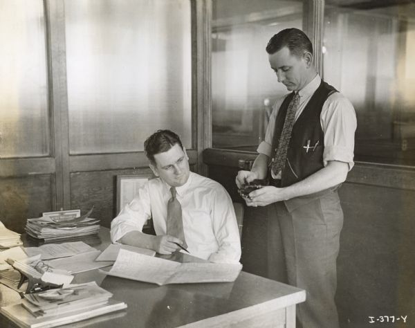 Charles W. Gilden sits at a desk in the Fort Wayne Works Engineering Department while Paul T. Brantingham stands next to him.