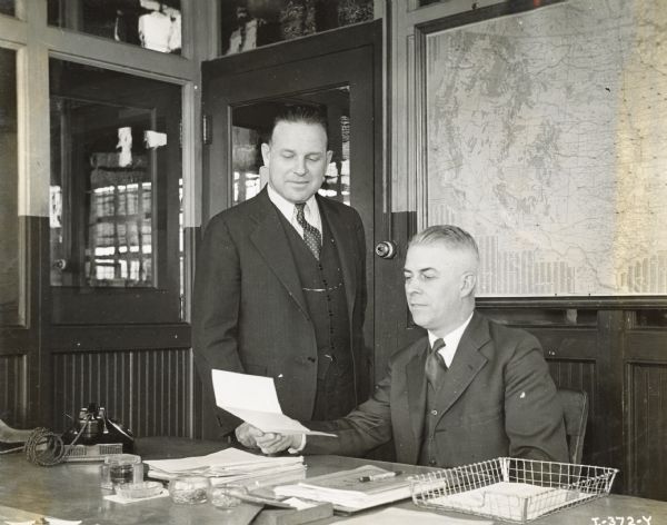 E.C. Lutz, superintendent of International Harvester's Fort Wayne Works, sits at a desk while C.M. Harrison, assistant superintendent, stands next to him.