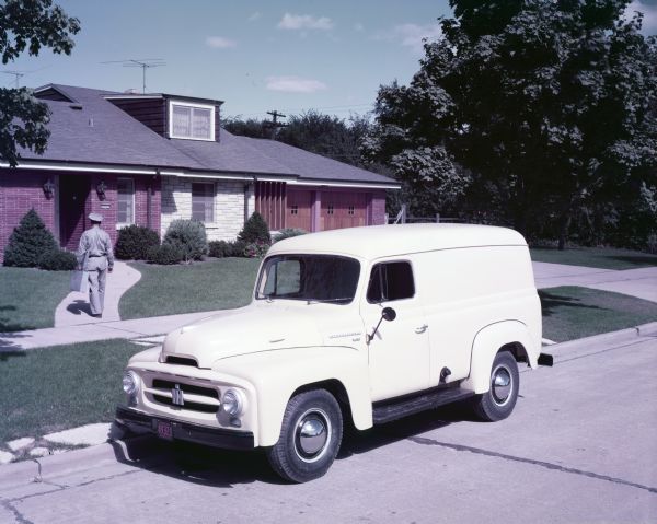 View of an International Model R-120 truck parked in front of a ranch-style home.  In the background, a uniformed man with a briefcase is walking toward the house's entrance.