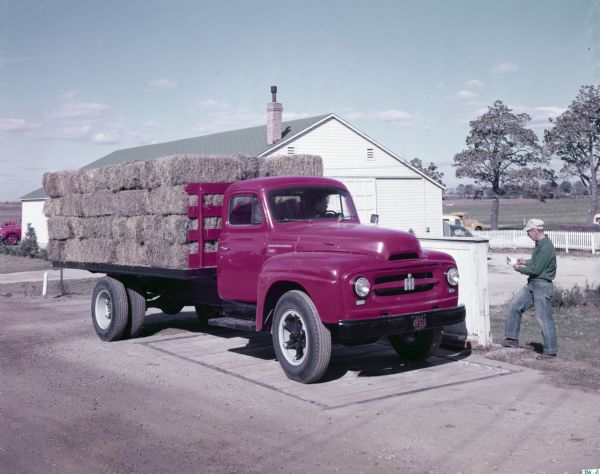 Color photograph of a man standing next to an International R-170 stake-body truck loaded with bales of hay.