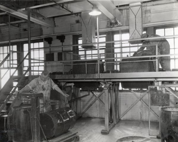 An factory worker in the Paint Department at McCormick Works discharges 100-gallon ball mills. Another man works in the background on a raised platform in front of windows.