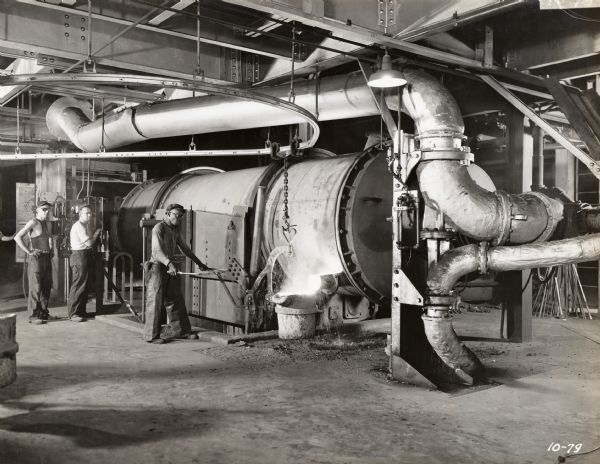 Factory workers at McCormick Works tap an 8-top Brackelsberg furnace. The original caption reads: "Control mechanism for rotating and tilting furnace, also primary and secondary air and pulverized coal firing equipment mounted on control board at left. Note cleanliness of installation."