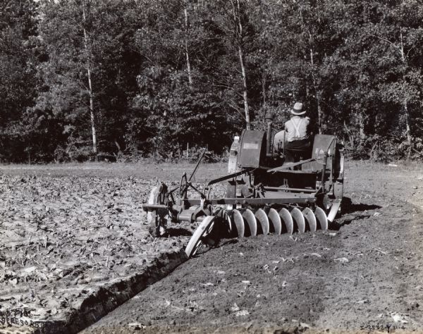 A farmer operates a 15-30 tractor with a 9-foot disk harrow plow through a field near New Hamilton, Ontario, in Canada. Trees are in the background.