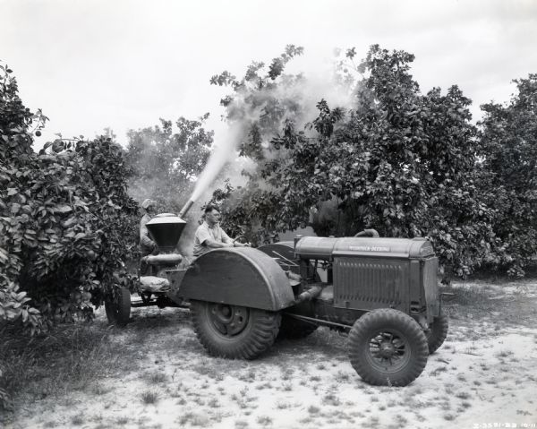 A man drives a McCormick-Deering 10-20 orchard and grove tractor with a 14-A disk and duster. Another man is dusting the trees with a pesticide.