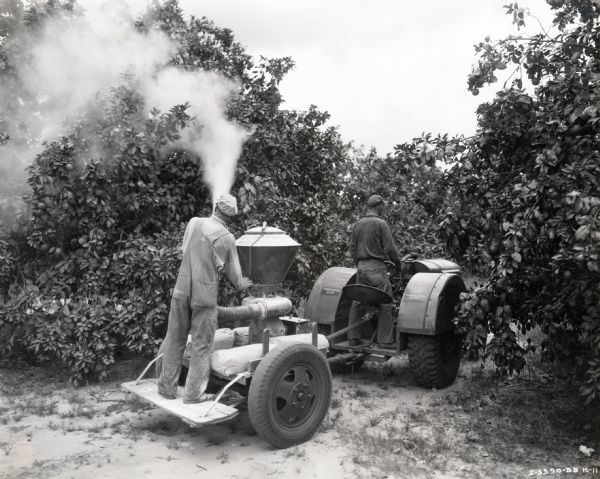 A 10-20 orchard and grove tractor pulls a McCormick-Deering 14-A disk and duster owned by Mackay Estates Ltd. A man is using the duster to apply pesticide to the trees.