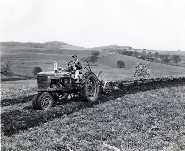 A farmer is using a Farmall H tractor to work in a hilly field. Shocks of corn are behind the tractor.