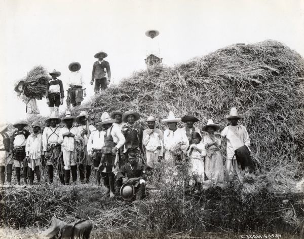 A group of individuals in native dress pose on and around a large haystack, possibly in Mexico.