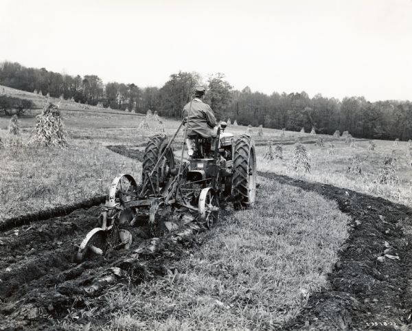 Hugh N. Brown is using a Farmall H and 2-bottom 14-inch plow to build diversion terraces. Shocks of corn are in the background. The original caption reads: "Hugh N. Brown of Route 1, Indiana, Pennsylvania, who recently purchased a 138-acre farm is shown building diversion terraces with his new Farmall-H and 2-bottom 14-inch plow on a hillside field.  Slope of the field varies from 10 to 12 percent and width of strips between the terraces is 80 to 100 feet.  After the terraces were finished they were seeded to grass and Mr. Brown said he planned to keep these diversion terraces permanently in sod.  Corn and oats and also some soybeans will be planted in alternate strips."