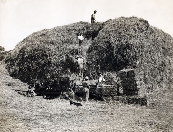 Australian farmers using an International kerosene engine and hay press to work with a large pile of hay.