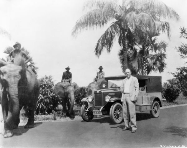 Clyde N. King standing next to an International Special Delivery truck at the "Mission in Buta" in the Belgian Congo. Three men are sitting on elephants nearby. King was part of an expedition sponsored by International Harvester to drive a truck across Africa.