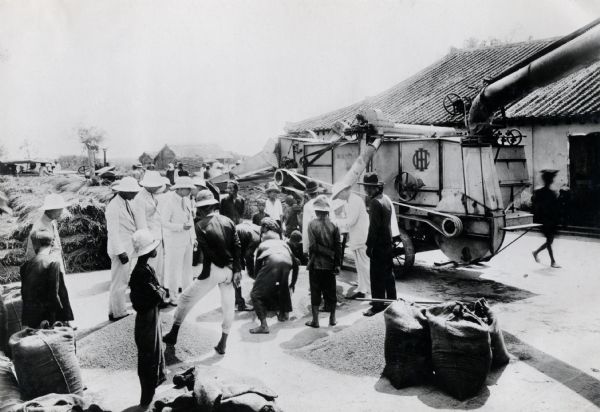 A group of people is gathered around an International Harvester stationary threshing machine. The original photographic caption reads: "Indo China."