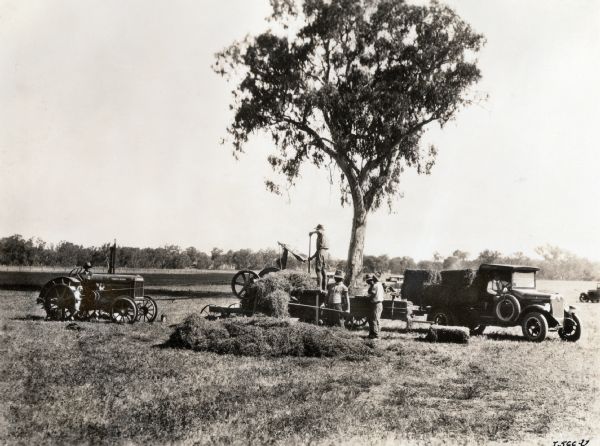 A group of farmers loads hay into a hay press on an Australian farm. The men are also using a McCormick-Deering 10-20(?) tractor and a pickup truck.