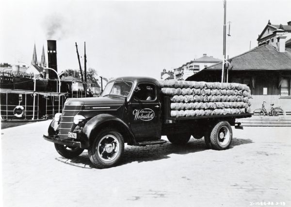 An International Model D-30 truck loaded with twine is parked near a body of water in Uppsala, Sweden. A large ship is in the background. The truck was owned by A.B. Wolrath & Co.