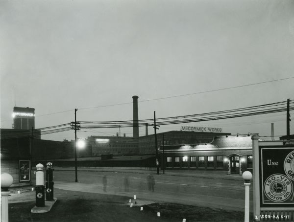 Exterior view of the McCormick Works factory at night. A tower with an illuminated sign reading "McCormick" is in the background. A faint image of a group of men, due to the long camera exposure, can be seen standing on the sidewalk.