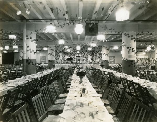 A hall, possibly at the McCormick Works Club House, decorated with vases of flowers, long tables laid with place settings, and chairs set up for a banquet or function. In the background is a piano, microphone, and music stands.