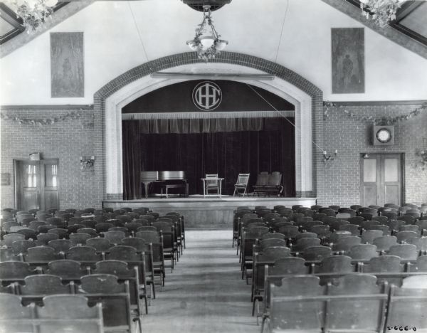 Rows of seats lead up to a arched stage outfitted with a piano and chairs at the McCormick Works Club House theater. The IHC logo is embroidered on the stage curtain. The Club House was designed for recreational use by factory employees.