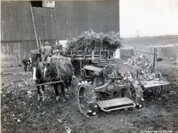A team of two horses with a wagon, and a Farmall Regular tractor, standing next to farmers as they are using a corn husker shredder outside a barn, possibly on the farm of Joseph Semperly.