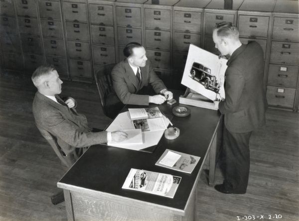 Elevated view of a man presenting an illustrated placard of an International Harvester motor truck to two other men sitting behind a desk. There is a large row of file cabinets behind them, and International truck advertising on the desk.