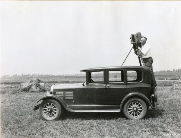 A company photographer sets up his camera on top of a car to take a photograph in a field.