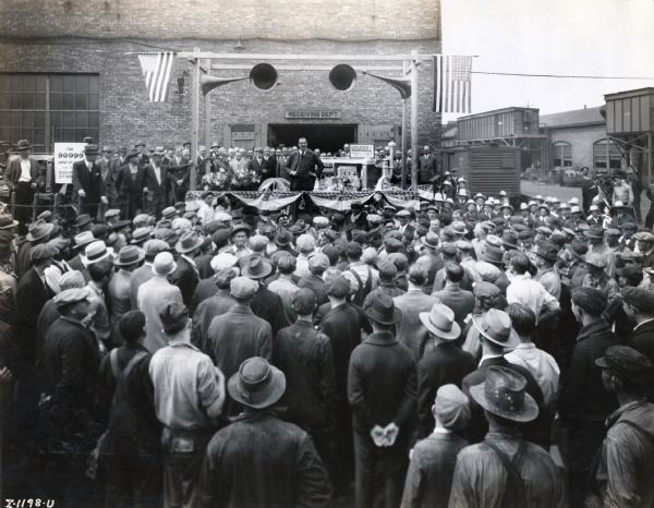Elevated view over crowd of people towards a man standing on a platform, possibly Cyrus McCormick III. He is speaking to a large group of people gathered outside the receiving department at the Farmall Works factory.  The sign on the platform reads: "100,000th Farmall, Farmall Works, April 12, 1930."