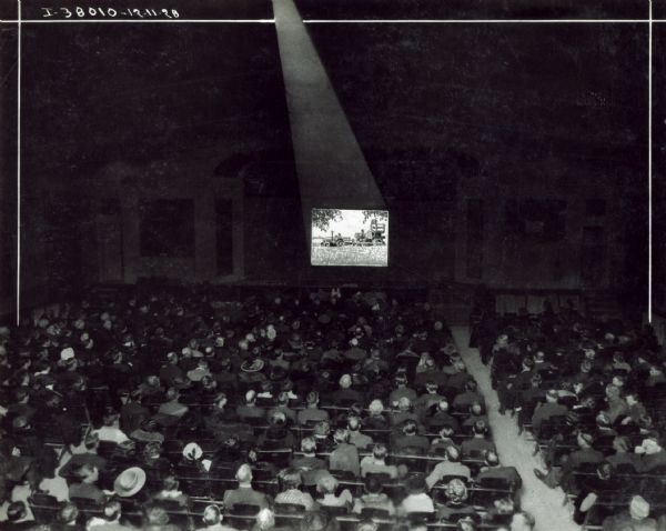 Rear view of a large audience gathered in a darkened theater to watch a motion picture, most likely an International Harvester advertising film. The image on the screen has been heavily retouched and shows a tractor and possibly a harvester-thresher (combine) at work in a field.