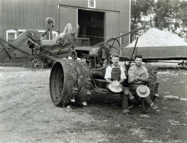 Two farmers posing for an outdoor portrait sitting on the back of a Farmall Regular tractor.