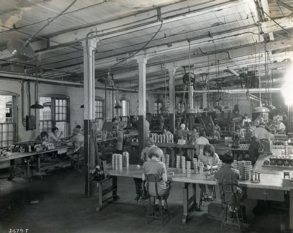Male and female factory workers at West Pullman Works sitting at tables in a large room to work with metal parts.