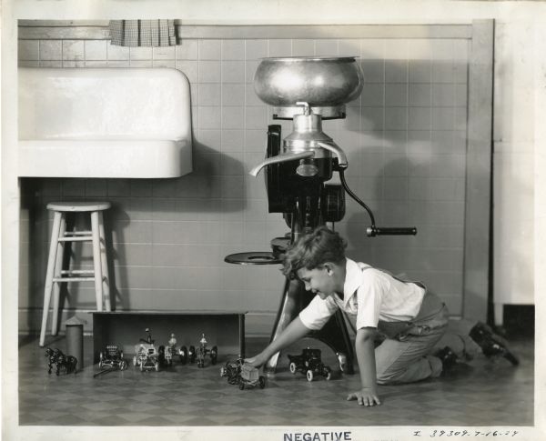 A boy is sitting on the floor near an International Harvester cream separator to play with toy versions of International Harvester machines, including Farmall tractors, a McCormick-Deering 10-20 tractor, an International truck, an all-steel thresher, a manure spreader and horses.