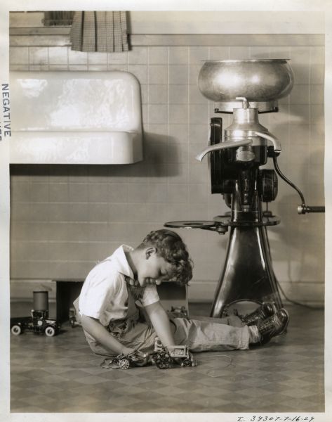 A young boy is sitting on the floor next to an International Harvester cream separator while playing with a toy tractor and plow. A toy International truck is in the background. A sink is on the wall on the left.