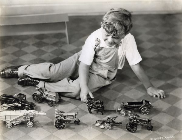 A boy is sitting on a tile floor to play with die-cast toys of International Harvester machines, including an all-steel thresher, Farmall tractors, McCormick-Deering 10-20 tractor, manure spreader, International truck, horses and a tractor plow.