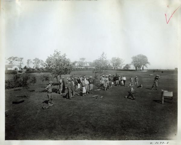 A group of men play golf at an International Harvester employee picnic. There are women and children watching the group.