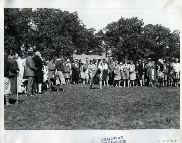 A group of people gather at a International Harvester employee picnic. A woman is standing at what appears to be a starting line, while a man stands by with a megaphone in his hand.