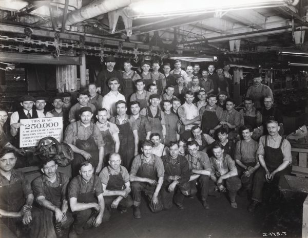 A group of factory workers poses for a photograph on the occasion of the 250,000th mower produced between 1926 and 1930, possibly at McCormick Works in Chicago. A man is holding a sign with the following text: "This is No. 250,000, a quarter of a million mowers over this progressive assembly chain from (?), 1926 to Feb. 2(?), 1930."