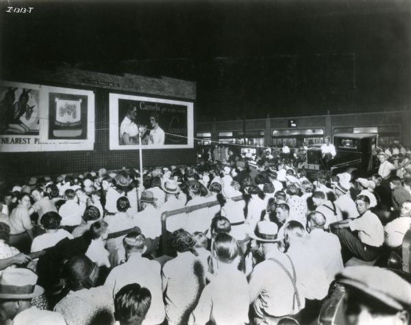 A group gathers to watch an International Harvester film projected from the bed of a pickup truck.