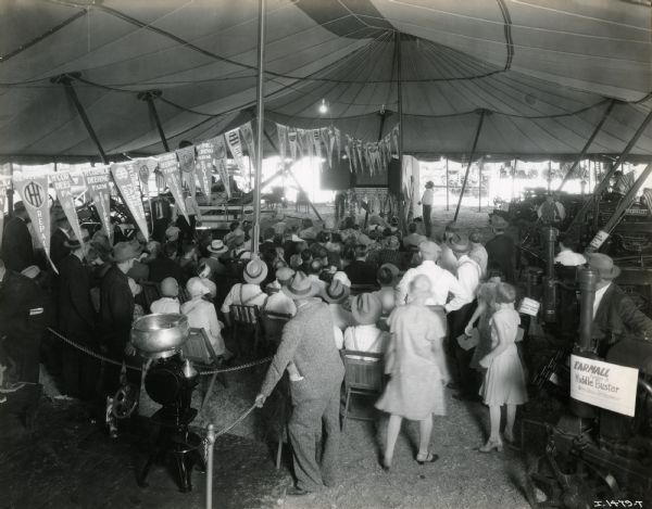 Elevated view of a group of Nebraska State fairgoers gathered in the International Harvester tent to watch a film. Advertising pennants hang from the tent's ceiling, and a cream separator and Farmall Middle Buster sit on display in the foreground.