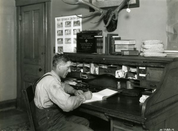 A man sitting at a desk writing on a tablet. A poster illustrated with cows is hanging on the wall in front of him, reading "World All-American Selections 192(?)." The man may be an International Harvester dealer.