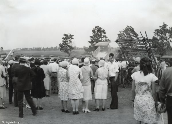 A crowd of people stands outdoors to look at agricultural machinery on display, including a hayloader and a farm truck.