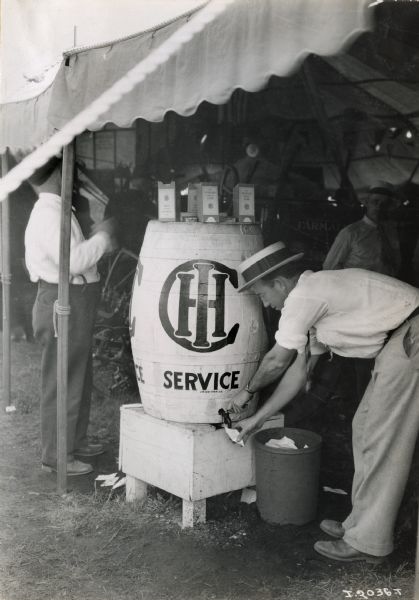 A man is stooping down to fill a paper cup with a beverage from a barrel dispenser under a tent at what appears to be a fair. The barrel is marked "IHC Service."