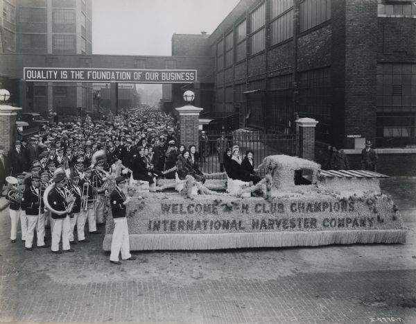 A group of young women at an International Harvester factory (most likely McCormick Works) ride on a parade float reading "Welcome 4-H Club Champions, International Harvester Company" while a marching band and spectators follow behind. A banner in the background reads: "Quality is the Foundation of our Business."