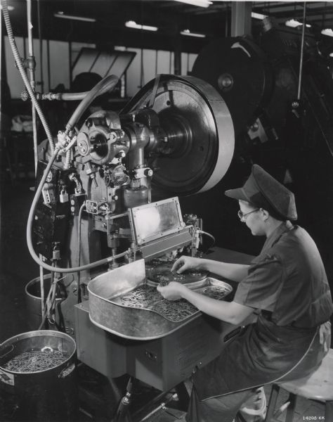 A female factory worker is sitting on a stool to work with metal parts in the ball bearing plant at West Pullman Works.