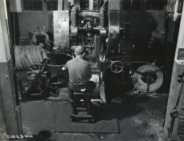 A factory worker is sitting with his back to the camera as he works at a machine at West Pullman Works. Coils of wire are lying on the floor.