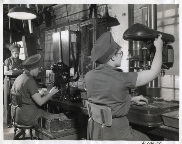 Female factory workers sit at machines at West Pullman Works, while another person is standing in the background.