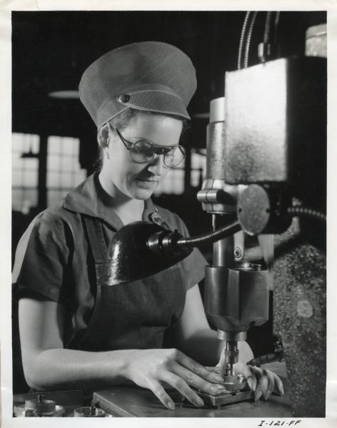 A female factory worker at West Pullman Works wears safety goggles to operate a "tapping machine." The original caption reads: "This photograph shows a woman employe (<i>sic</i>) of the Chicago West Pullman Works operating a tapping machine. She wears the type of safety cap worn by all women employes (<i>sic</i>) of the Company who work on machines that have revolving spindles. She also wears safety goggles which are worn by all production employes (<i>sic</i>) of the Company for protection to her eyes."
