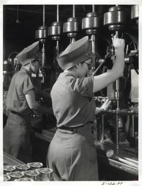 Factory workers at West Pullman Works operate a multiple drill machine. The original caption reads: "A side view of women employes (<i>sic</i>) of the Chicago West Pullman Works of the International Harvester Company shown operating a multiple drill machine. They wear safety caps to avoid their hair becoming entangled in any moving parts of the machine, and safety goggles for the protection of their eyes."