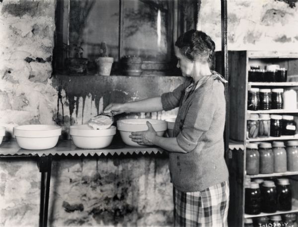 Mrs. L.A. Hawkins skimming cream from a bowl in a basement using an "old-fashioned" tin skimmer. Shelves of canned goods are in the background.