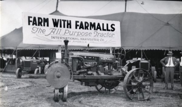 A Farmall Regular tractor stands on outdoor display near a tent at the Mid-South Fair. A sign reading: "Farm With Farmalls; The All-Purpose Tractor; International Harvester Co. of America" is displayed on the tent. The tractor has a saw attachment.
